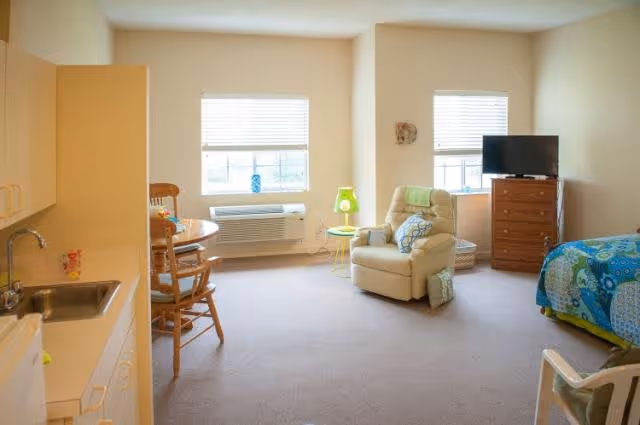 A bright and cozy senior living room with beige walls and carpet. The room features a single bed with a colorful blue and green quilt, a beige recliner chair with a green pillow and a small green side table with a lamp. There is a wooden dresser with a flat-screen TV on top, two windows with blinds letting in natural light, a round wooden dining table with chairs, and a kitchenette area with a sink and cabinets.