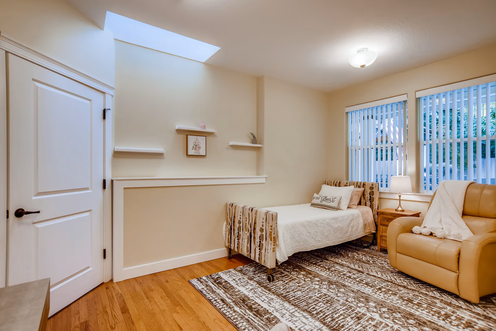 A cozy bedroom with a single bed featuring a patterned headboard and footboard, a white quilt, and a pillow that says 'Our Guest'. Next to the bed is a wooden nightstand with a lamp. There is a comfortable tan recliner chair with a white throw blanket draped over it. The room has hardwood floors with a large patterned area rug, beige walls, a white door, and two windows with vertical blinds letting in natural light. A skylight is visible on the ceiling, and there are small decorative shelves on the wall above the bed.