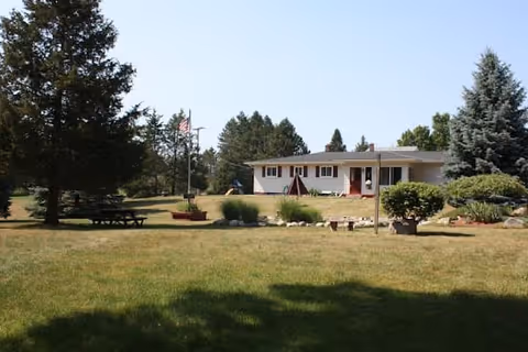 A single-story white building with a red door surrounded by a large grassy yard with trees and shrubs. There is an American flag on a flagpole near a picnic table on the left side of the yard. The sky is clear and blue.