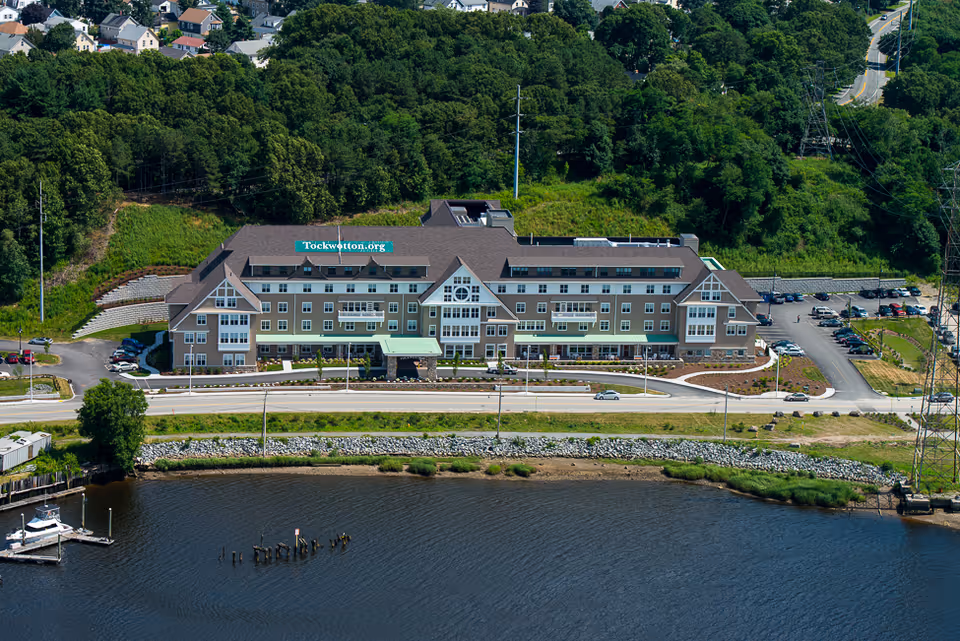 Aerial view of Tockwotton on the Waterfront, a large multi-story senior living facility situated near a body of water with a dock and surrounded by greenery and residential houses in the background.