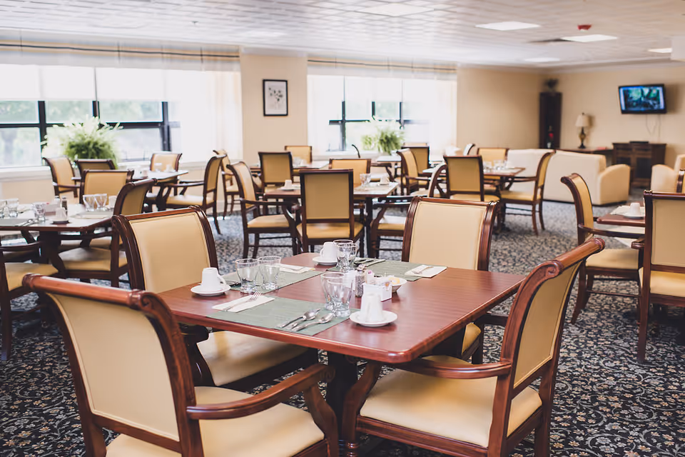 A bright dining room with multiple wooden tables and beige cushioned chairs arranged neatly. Each table is set with placemats, cups, glasses, and utensils. Large windows with white blinds allow natural light to fill the room, and there are plants on the windowsills. In the background, there is a television mounted on the wall and a lamp on a wooden cabinet.