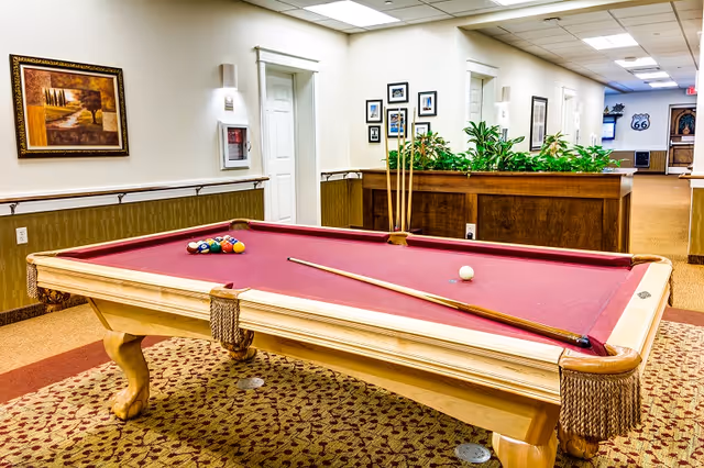 A red-felt billiards table in a well-lit interior common area of a senior living facility with plants, framed art, and hallways in the background.