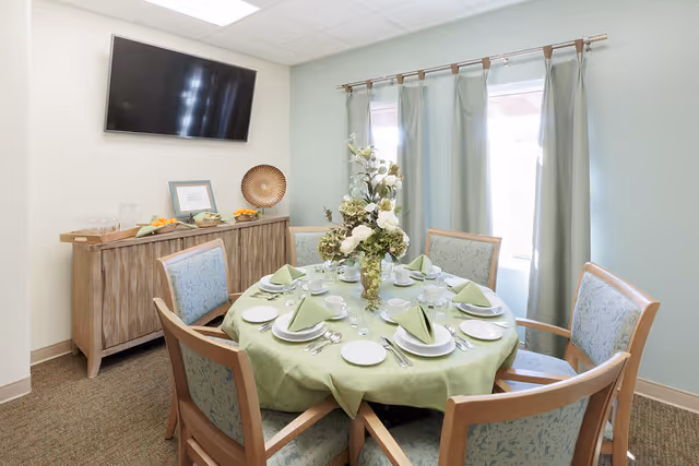 A small dining room with a round table covered with a light green tablecloth set for six people with plates, cups, utensils, and folded green napkins. A floral centerpiece is in the middle of the table. There are six wooden chairs with light blue patterned cushions around the table. Behind the table is a wooden sideboard with a tray of glasses, a framed picture, and a decorative plate. A flat-screen TV is mounted on the wall above the sideboard. Light blue curtains cover a window on the right side of the room.