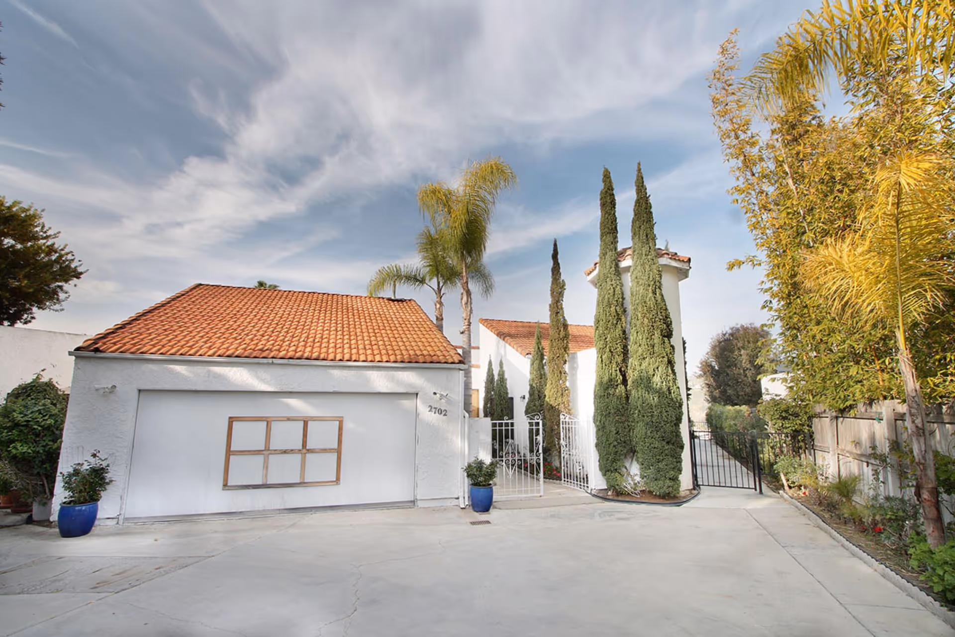 Front exterior of a white stucco building with a red tile roof, garage door, tall cypress and palm trees, and a gated driveway.
