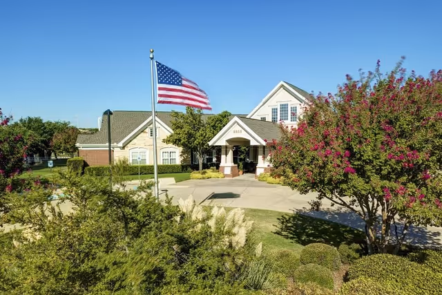 Front entrance of a senior living building with an American flag, landscaped driveway, and flowering trees.
