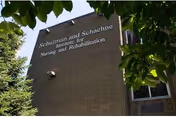 Exterior view of a brick building with the sign 'Schulman and Schachne Institute for Nursing and Rehabilitation' on the wall, partially framed by green tree leaves.