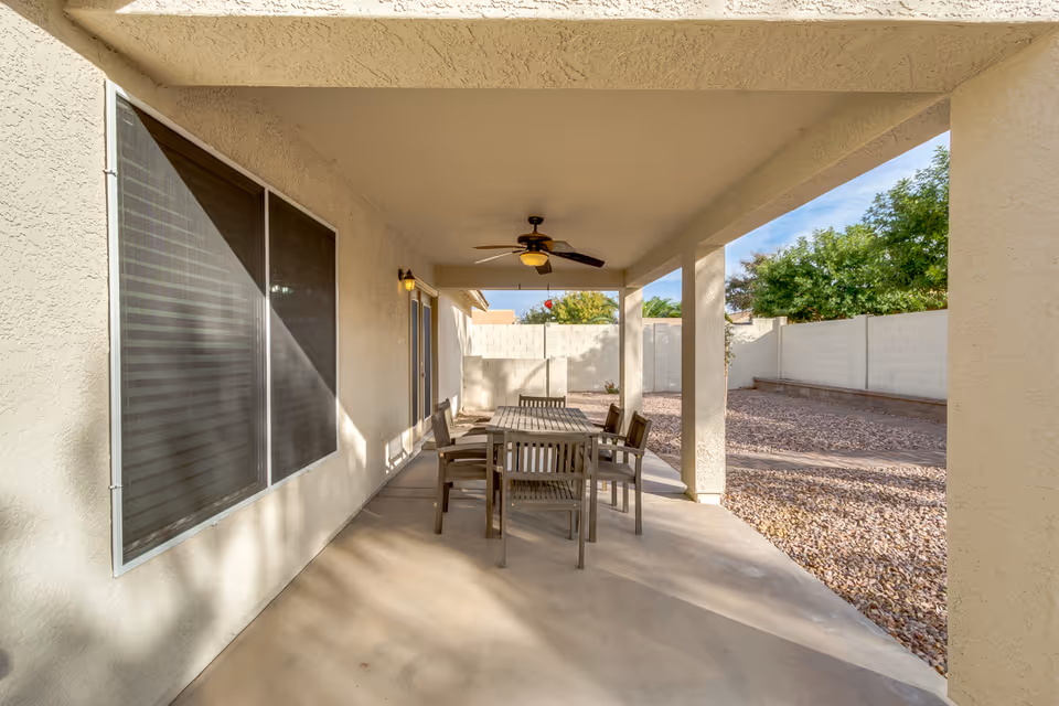 Covered outdoor patio area with a ceiling fan and a wooden table surrounded by six chairs. The patio is adjacent to a beige stucco wall with a large window and a door. Beyond the patio is a yard with gravel ground cover and a white block wall fence, with some green trees visible in the background.