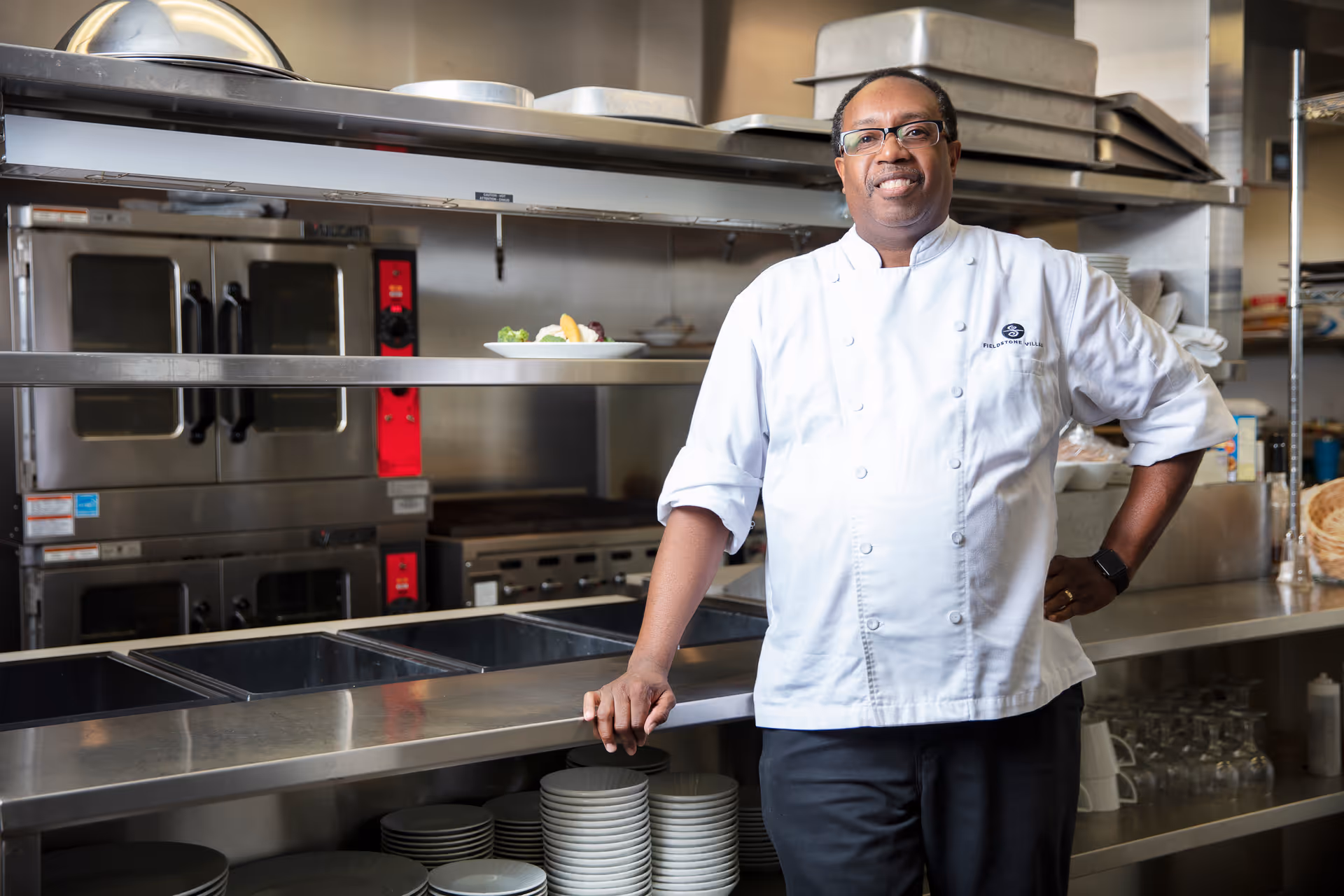 A chef in a white uniform stands in a stainless-steel commercial kitchen by shelves of plates and ovens.