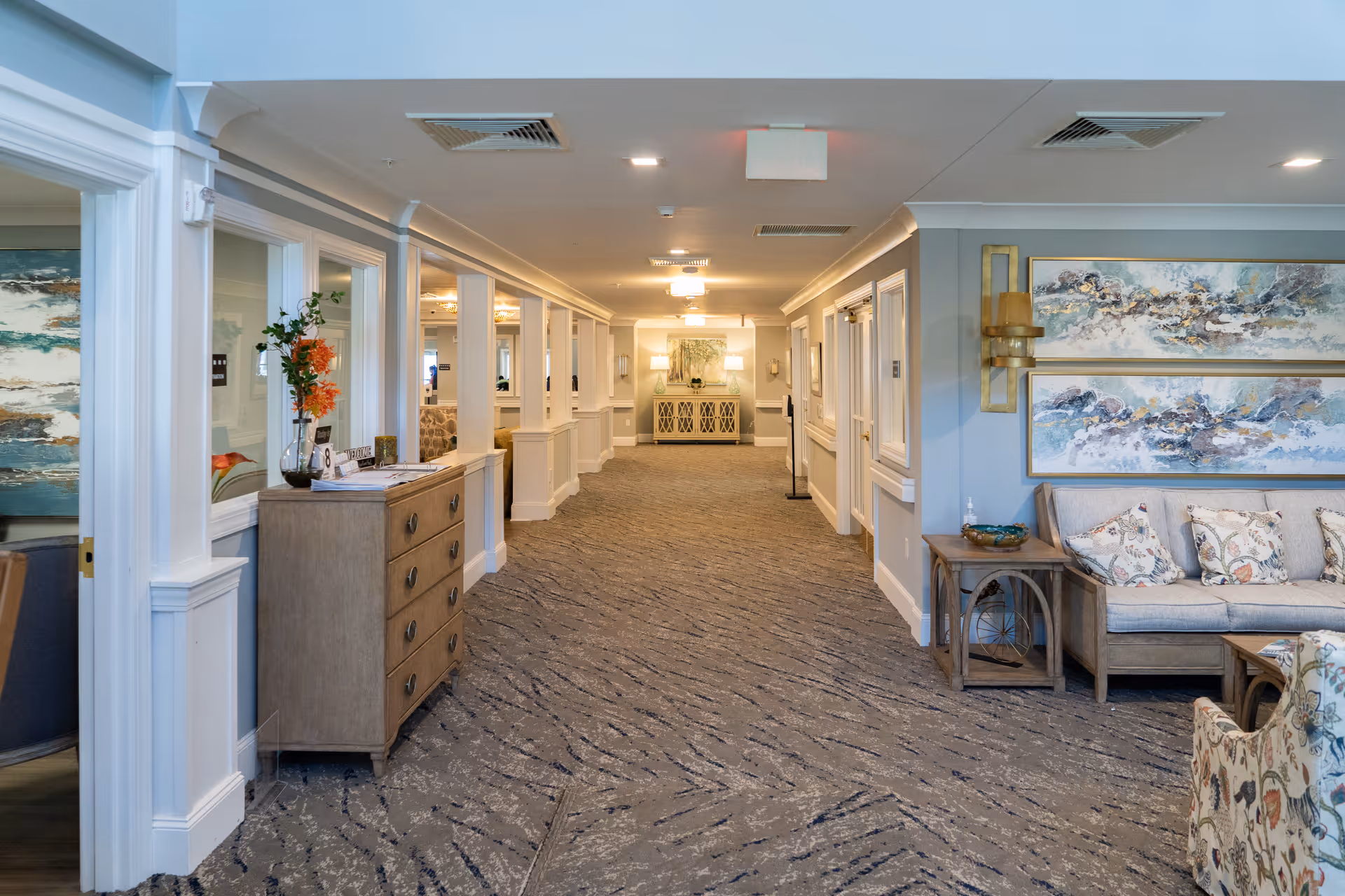A well-lit hallway in a senior living facility with patterned carpet, light gray walls, and white trim. On the right side, there is a seating area with a beige couch, floral pillows, a side table, and two abstract paintings on the wall. On the left side, there is a wooden chest of drawers with a vase of flowers and some papers on top. The hallway extends toward a cabinet with lamps and a framed artwork at the far end.