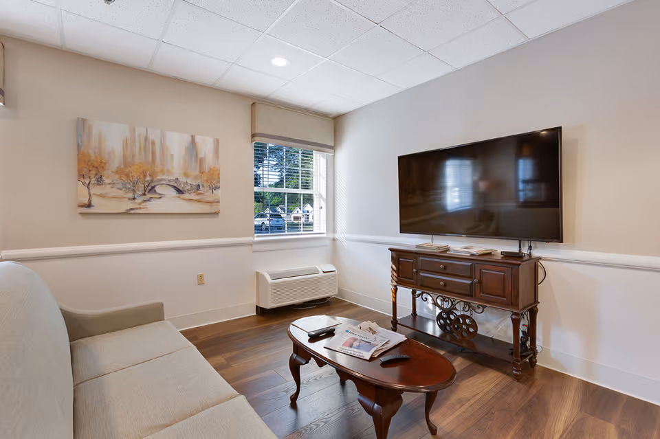 A cozy living room with a beige sofa on the left, a wooden coffee table with newspapers and remote controls in the center, and a large flat-screen TV mounted on the wall above a wooden console on the right. A window with blinds lets in natural light, and a painting of a cityscape with autumn trees hangs on the wall above the sofa.