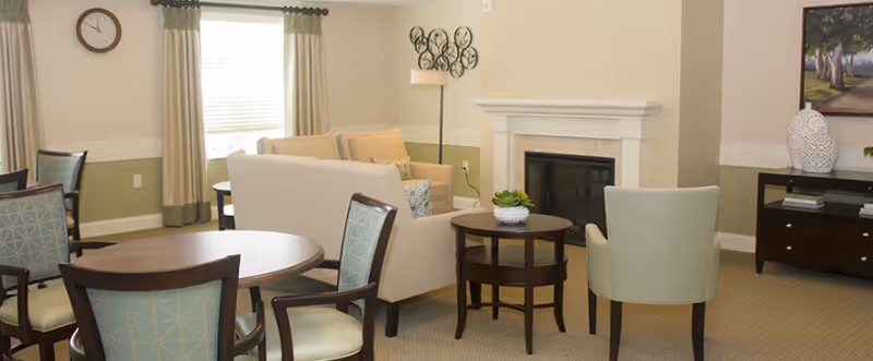 A cozy living room area with a beige sofa, two light green armchairs, a round wooden coffee table with a small plant, and a fireplace. There is a round dining table with four chairs in the foreground, a wall clock, and a window with beige curtains. A dark wooden cabinet with decorative items and a painting is visible on the right side.