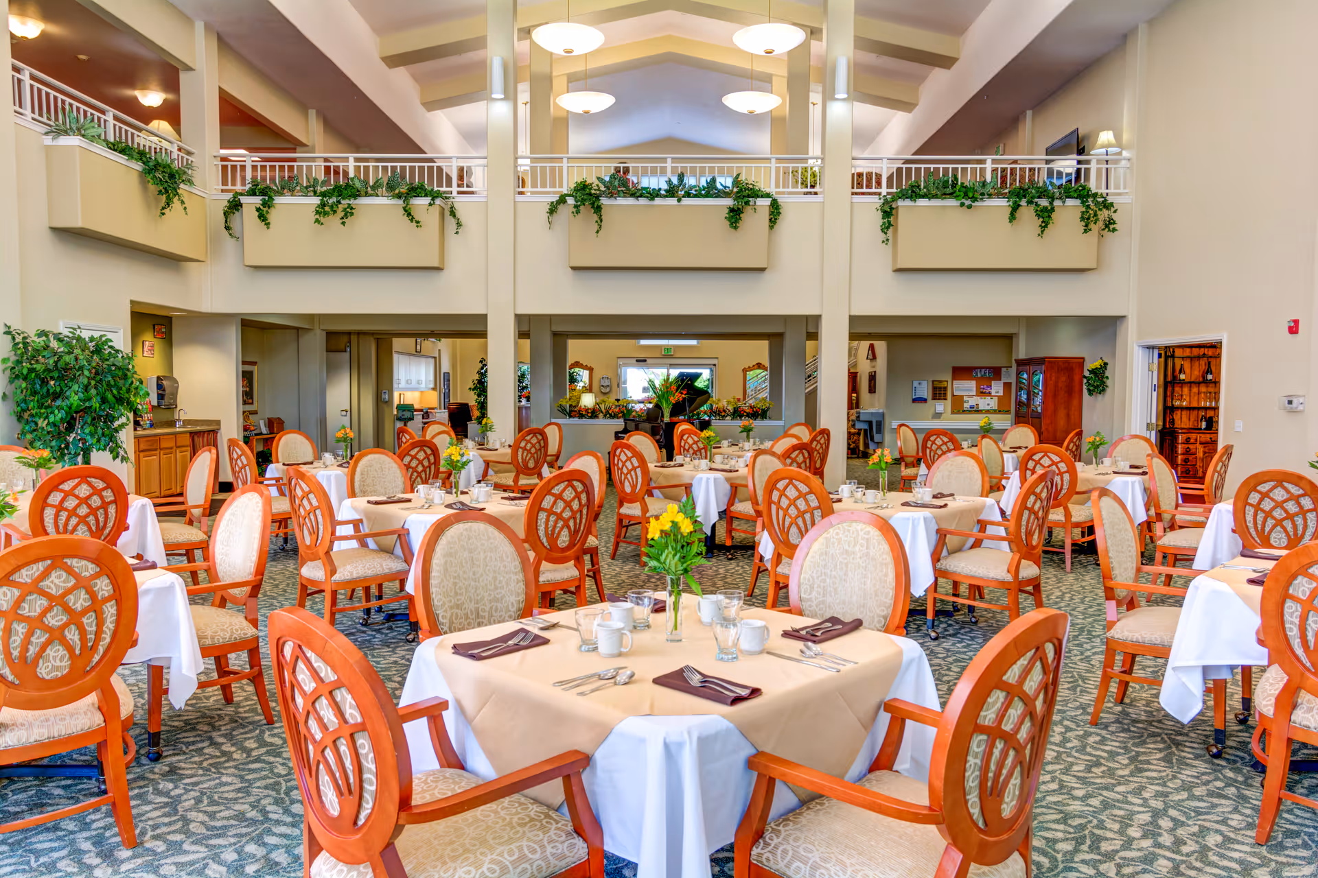 A spacious and well-lit dining room with multiple round tables covered with beige and white tablecloths. Each table is set with glasses, cups, silverware, and napkins. The chairs have wooden frames with patterned upholstery. The room features high ceilings, large planters with greenery on the upper level, and a carpeted floor with a leaf pattern. There are plants and decorative elements around the room, creating a welcoming atmosphere.