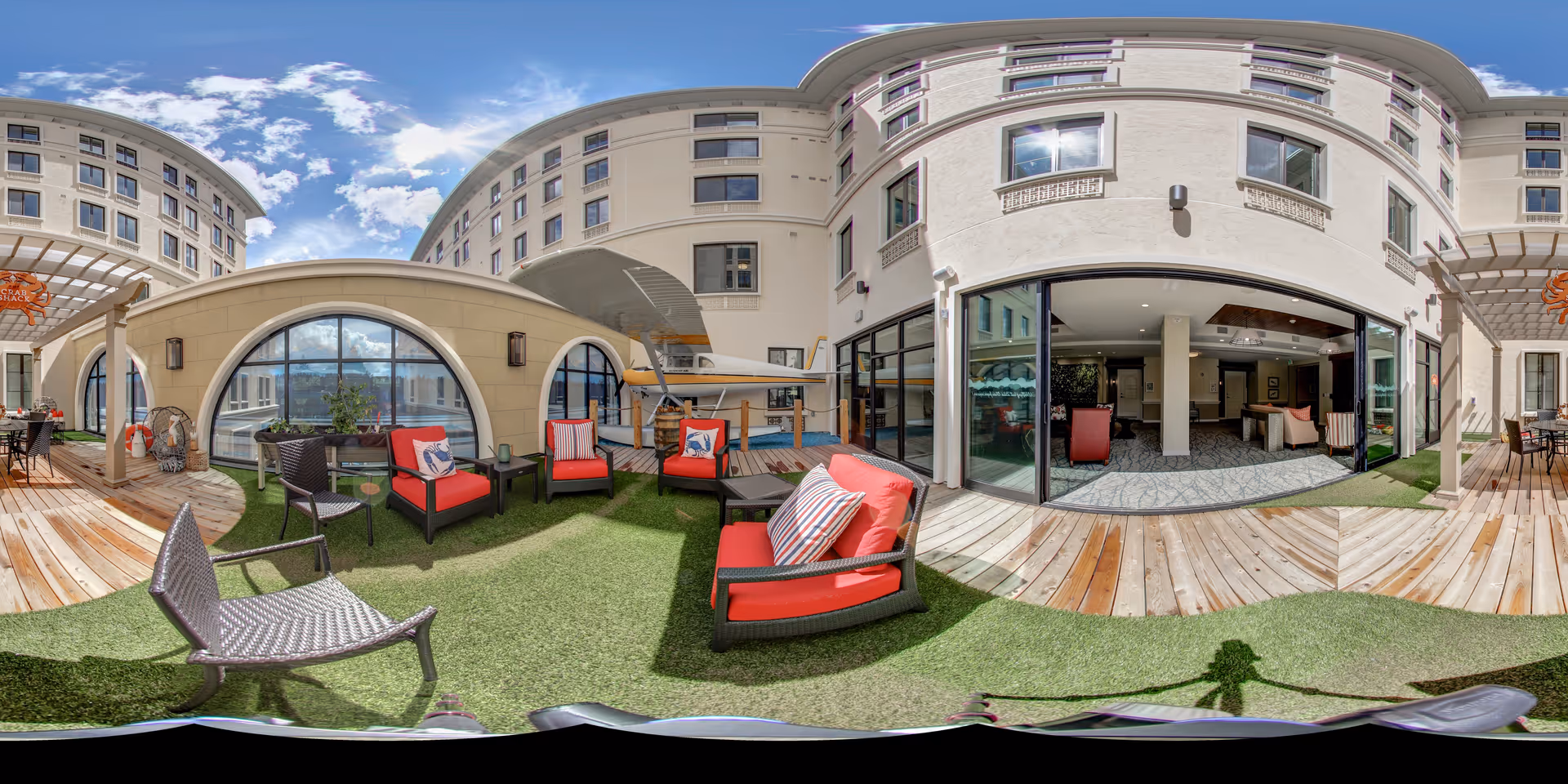 Outdoor patio area at Aegis Living Bellevue Overlake with red cushioned chairs, black wicker chairs, small tables, and artificial grass. The patio is surrounded by a multi-story building with large windows and sliding glass doors leading to an interior lounge area. The sky is partly cloudy with sunlight illuminating the scene.