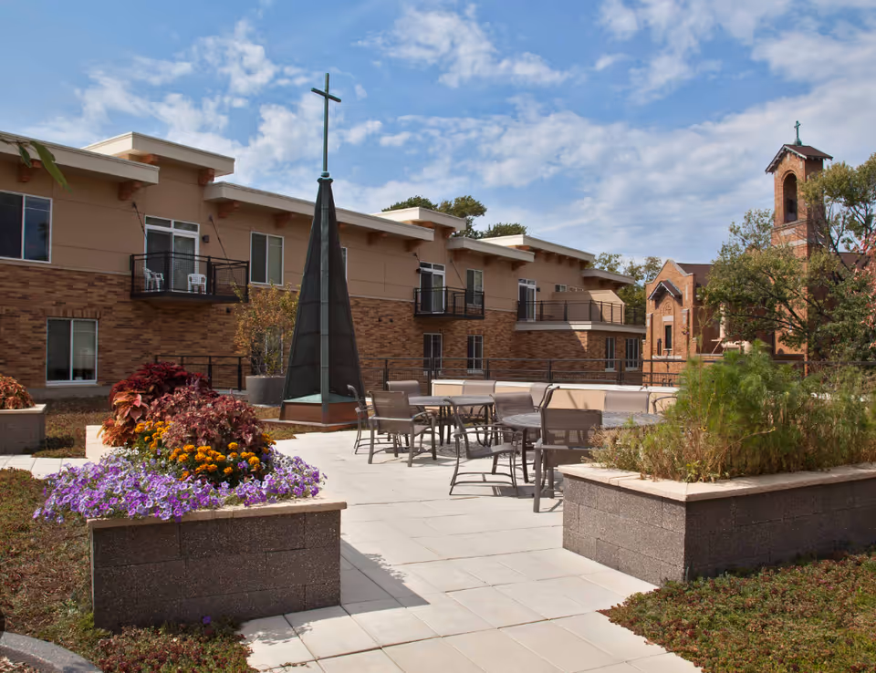 Outdoor patio area at Carondelet Village with tables and chairs surrounded by raised flower beds. A tall cross structure is centrally located, and a brick building with balconies is in the background. Another building with a bell tower and cross is visible to the right under a partly cloudy sky.