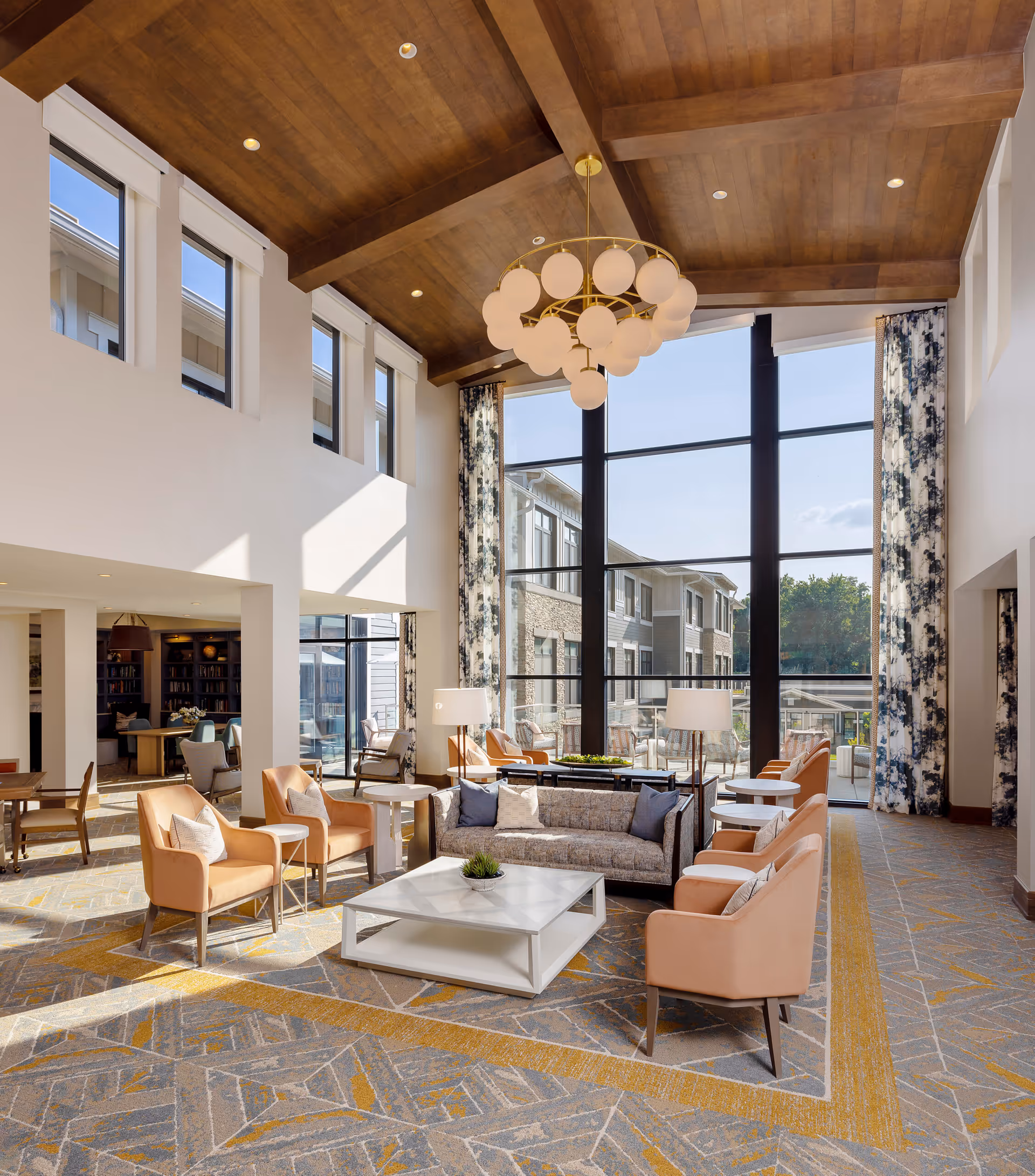 Spacious sunlit communal lounge with a high wood-beamed ceiling, floor-to-ceiling windows, a modern chandelier, and sofas and armchairs arranged around a central coffee table.