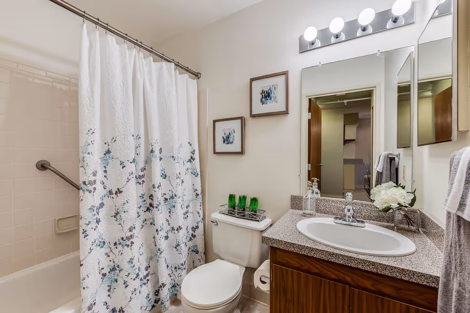 A clean bathroom featuring a bathtub with a white shower curtain decorated with blue and purple floral patterns. There is a white toilet with a decorative tray holding green glass items on top. Above the toilet, two framed abstract art pieces hang on the wall. A countertop with a sink, soap dispenser, and a small vase with white flowers is visible. A large mirror with lights above it reflects part of the room and an open door.