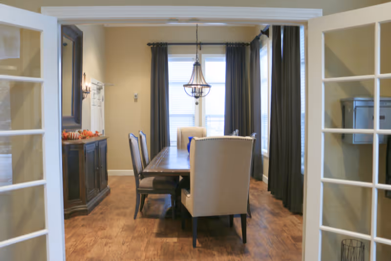 A dining room with a wooden table surrounded by six chairs, beige walls, dark curtains on the windows, a chandelier hanging above the table, and a wooden sideboard with decorative items on the left side.