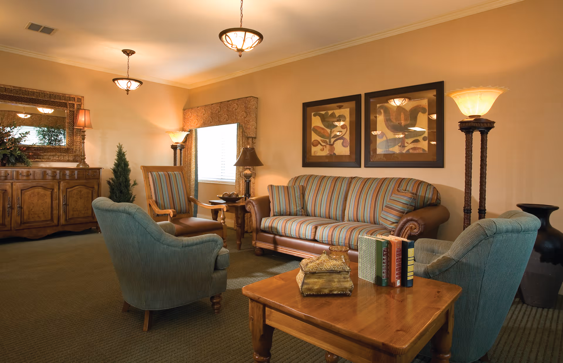 A cozy living room area with a striped sofa, two blue armchairs, a wooden coffee table with books and a decorative box, a wooden sideboard with a large mirror above it, two framed abstract paintings on the wall, and several lamps providing warm lighting.
