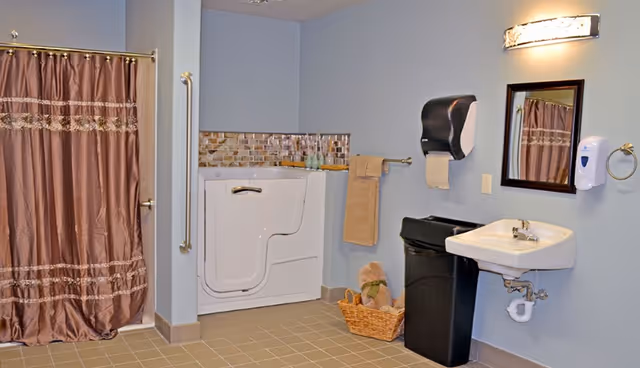 A spacious bathroom with a walk-in bathtub, a brown shower curtain, a wall-mounted sink with a mirror above it, a black trash bin, a paper towel dispenser, a soap dispenser, a towel rack with a beige towel, and a basket with rolled towels on the floor.