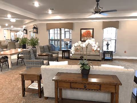 A spacious and well-lit living room area in a senior living facility featuring beige sofas, plaid armchairs, wooden side tables, a wooden console table with a white orchid plant, large windows with beige valances, and framed artwork on the wall.