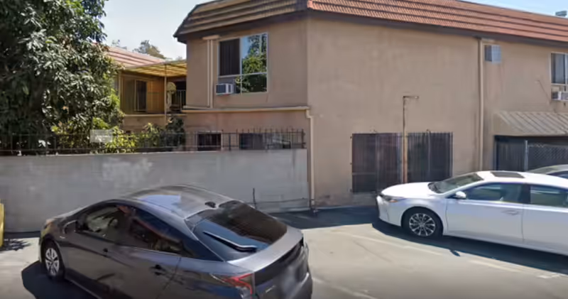 Parking area outside a beige two-story building with barred windows and air conditioning units. Two cars, one gray and one white, are parked in the lot. There is a concrete wall with greenery behind it on the left side.