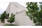Exterior view of a beige multi-story building with the name 'Regency' displayed near the top corner. There are small trees and shrubs planted along the sidewalk in front of the building.