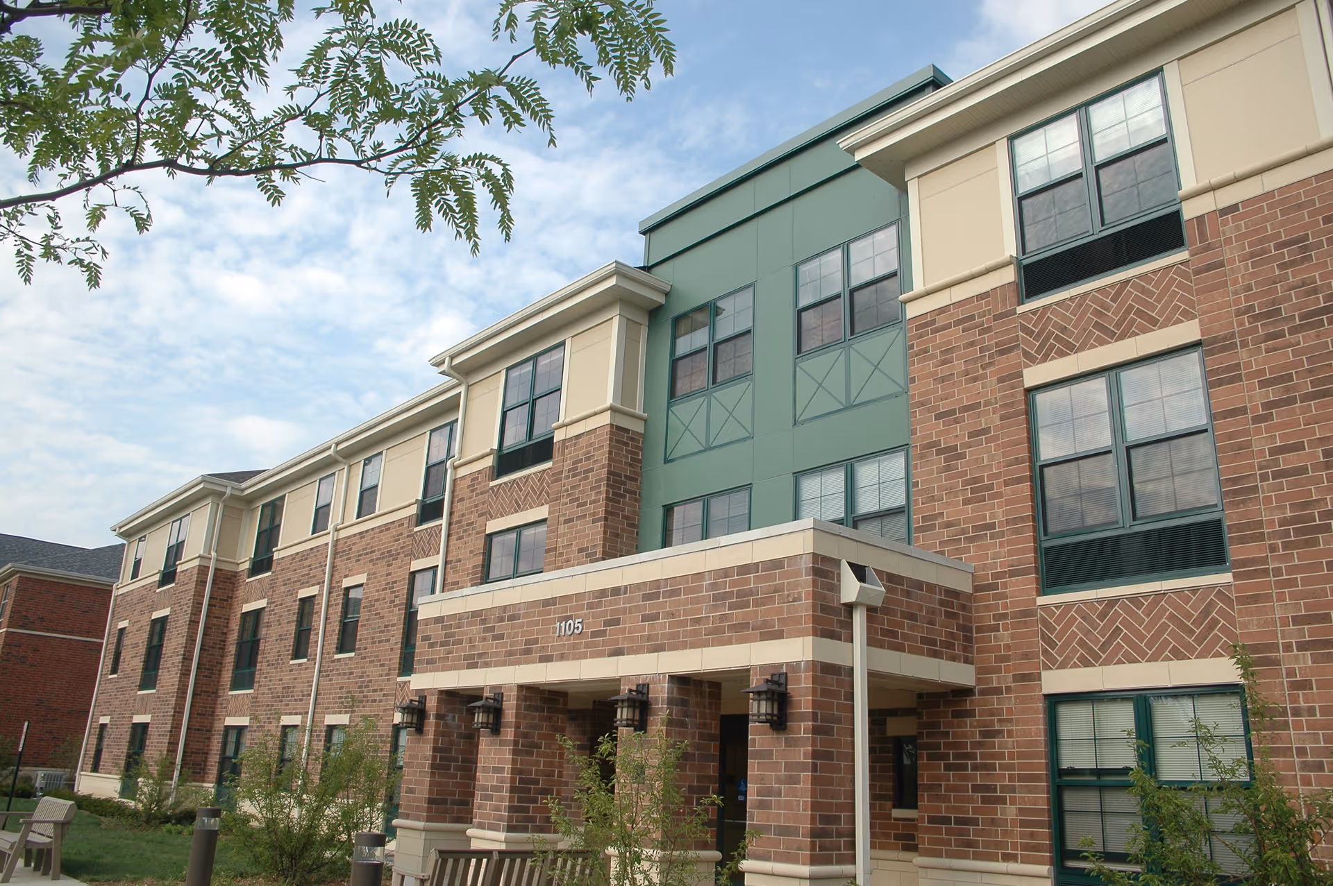 Exterior view of a three-story brick building with green and beige accents, featuring multiple windows and an entrance with a covered porch. There are small bushes and benches in front of the building under a partly cloudy sky.