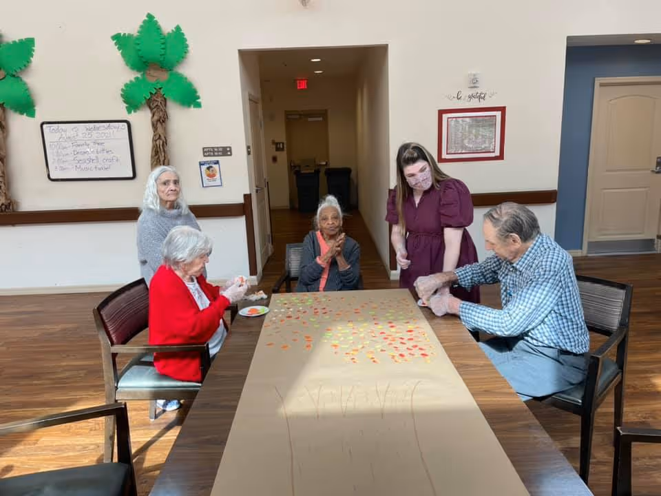 Four elderly individuals and a caregiver wearing a mask are gathered around a table in a senior living facility. They are engaged in a craft activity involving a large sheet of paper with a tree outline and colorful leaf shapes. The room has wooden flooring, decorative palm trees on the wall, and a whiteboard with a schedule.