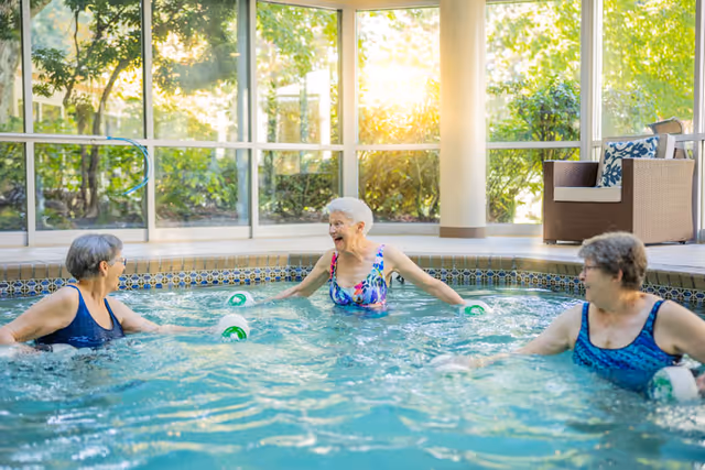 Three elderly women in swimsuits enjoying a water exercise session in an indoor pool with large windows showing greenery outside. One woman in the center is smiling and engaging with the others.