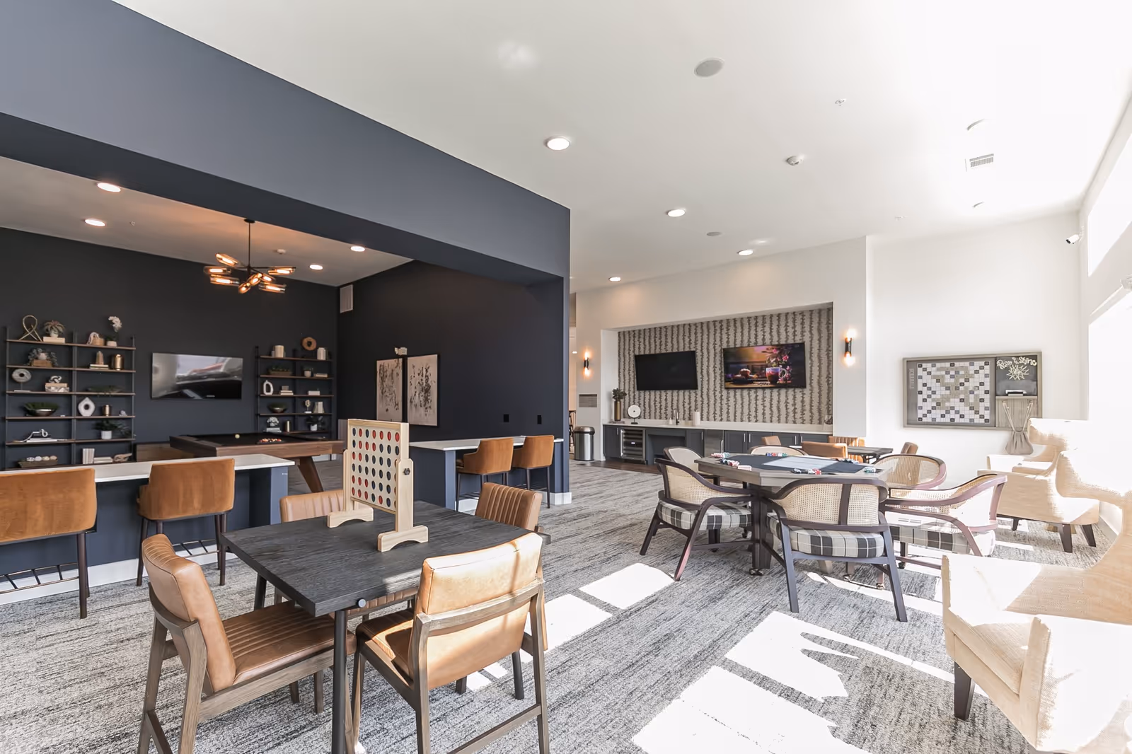Bright communal activity room with multiple tables and chairs, a bar counter with stools, games and wall-mounted TVs.