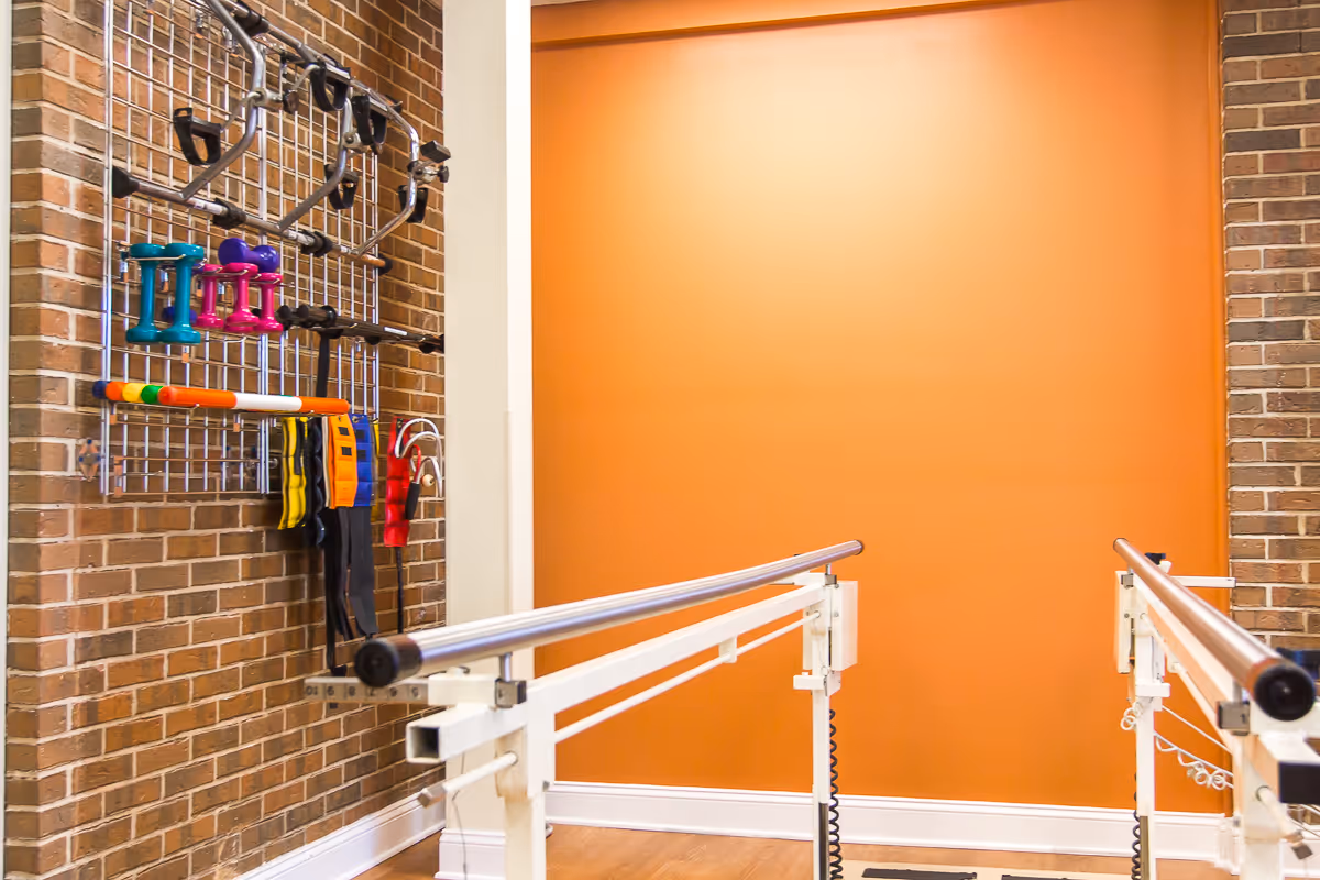 Physical therapy room with parallel bars for walking exercises and a wall-mounted rack holding various exercise equipment including dumbbells, resistance bands, and other fitness tools. The room has an orange accent wall and exposed brick walls.