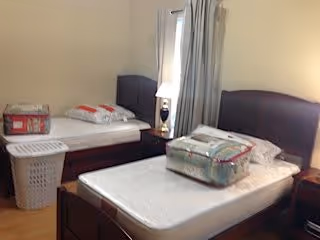 A bedroom with two single beds, each with a dark wooden headboard. One bed has a pillow and a folded blanket on it, while the other bed has a packaged bedding set placed on the mattress. There is a small nightstand with a lamp between the beds, and a laundry basket is positioned near the foot of one bed. Light-colored curtains cover a window in the background.