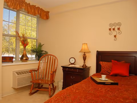 Well-lit bedroom with red bedding, a nightstand and lamp, and a wooden rocking chair by a window.