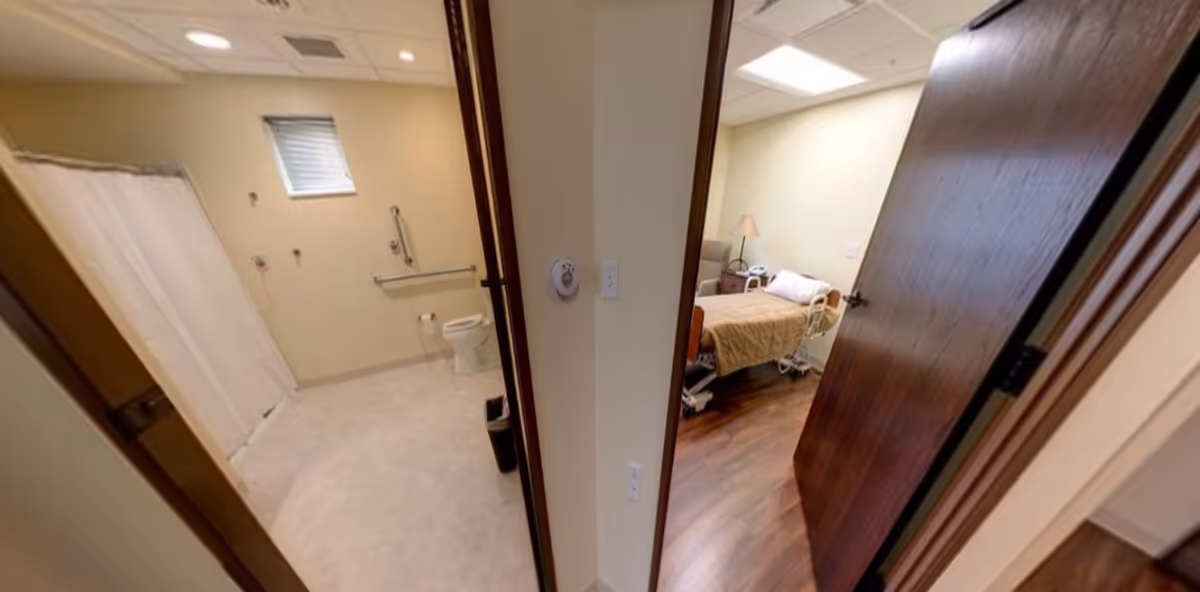 Accessible bathroom with shower and toilet on the left and a furnished bedroom with a hospital-style bed on the right.