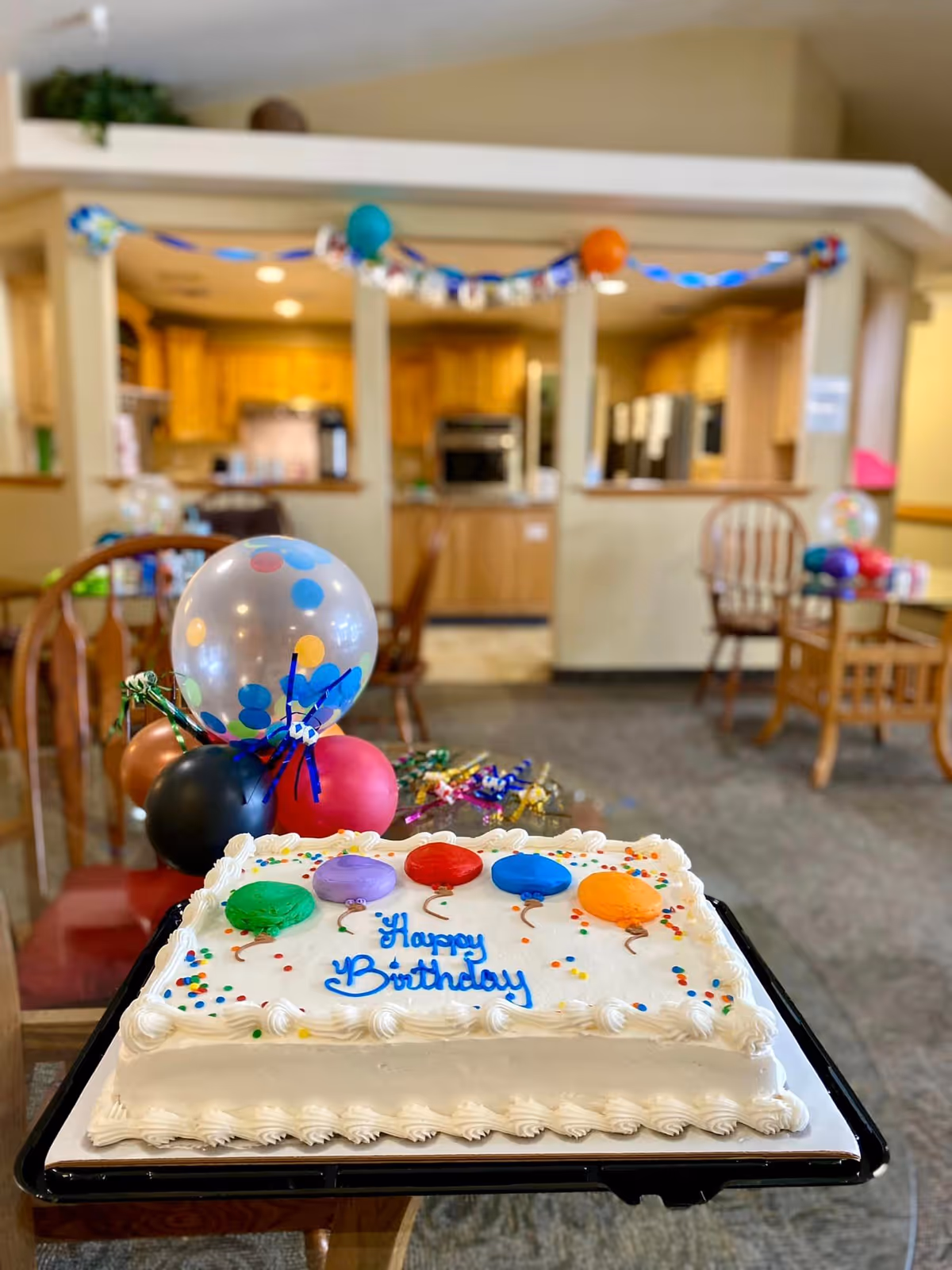 A decorated birthday cake with colorful balloon designs and the words 'Happy Birthday' written in blue icing is placed on a table in a room. Behind the cake, there are colorful balloons and party decorations. The background shows a kitchen area with wooden cabinets and appliances, and some chairs and tables are visible in the room.