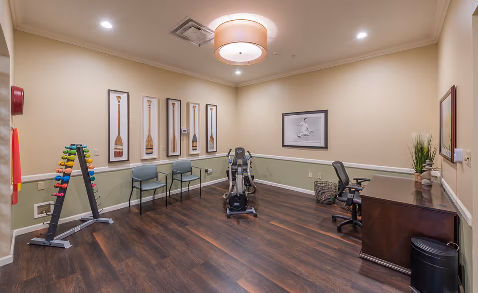 A small exercise room with wooden flooring and beige walls. The room contains a rack of colorful dumbbells, two chairs, an exercise bike, a desk with an office chair, and decorative framed pictures on the walls. There is a ceiling light fixture and some plants on the desk.