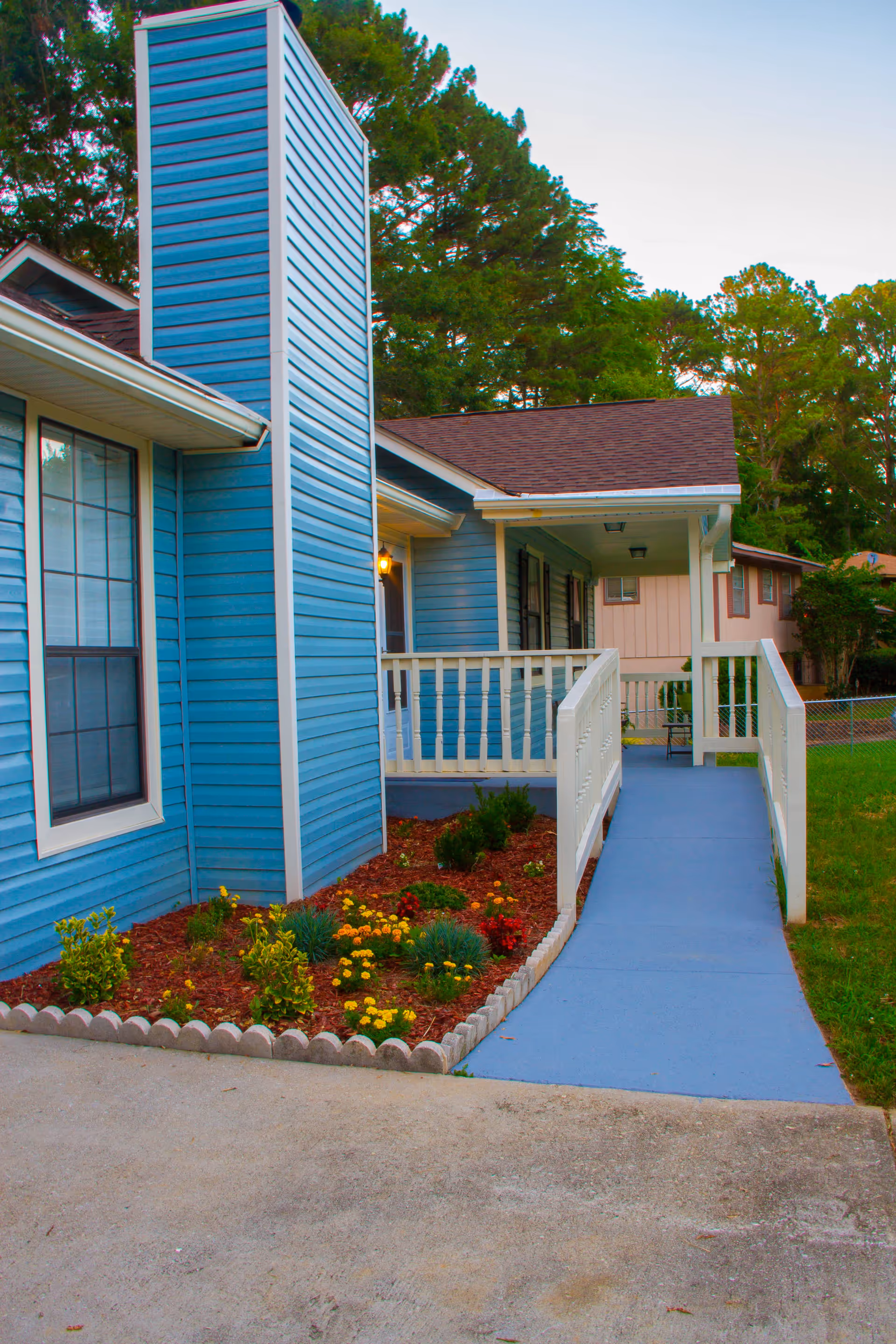 Exterior view of a blue residential building with a ramp leading to a covered porch. There is a small garden bed with colorful flowers and shrubs along the side of the building. Trees and another house are visible in the background.