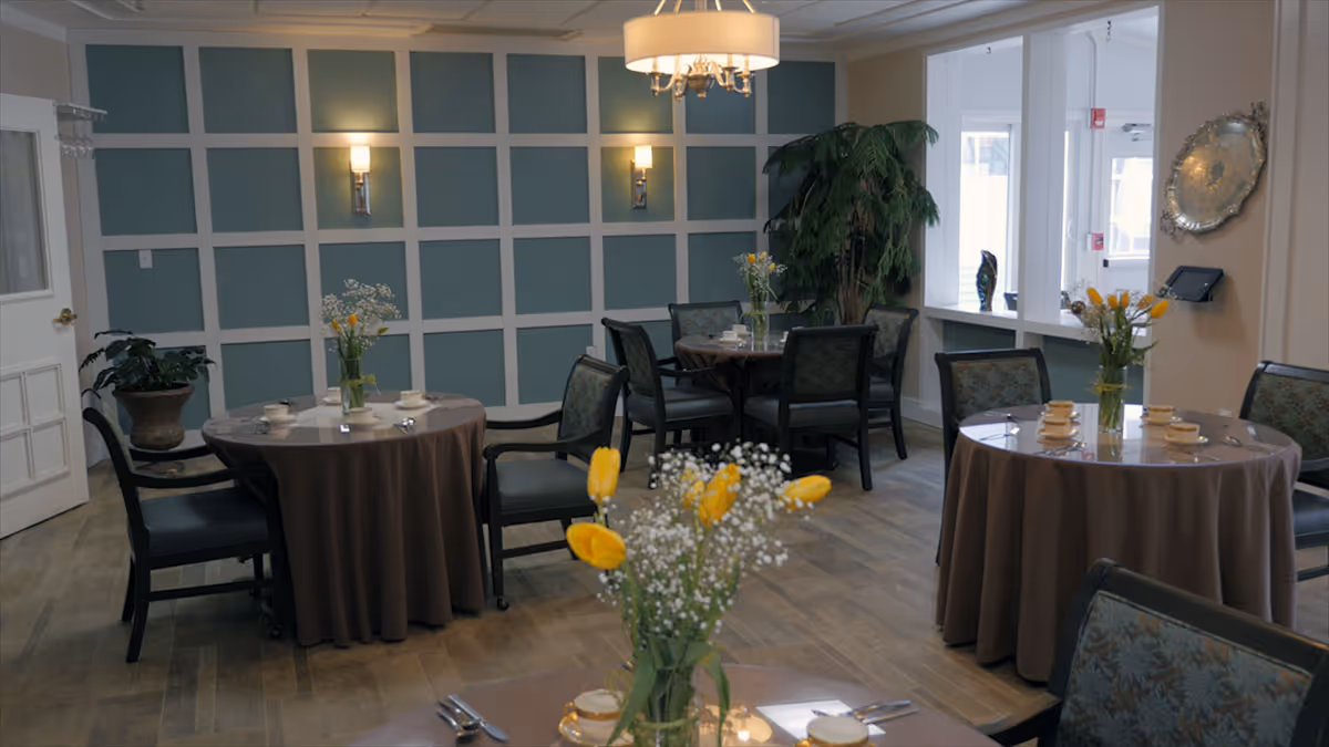 A dining room with round tables covered in brown tablecloths, each set with cups, saucers, and silverware. Yellow tulips and white baby's breath flowers in glass vases decorate the tables. The room has a blue and white paneled wall, wall sconces, a chandelier, and a large potted plant near a windowed partition.