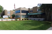 Exterior view of a two-story senior living facility building with large windows, a green awning over the entrance, a well-maintained lawn, and an American flag on a flagpole near the entrance.