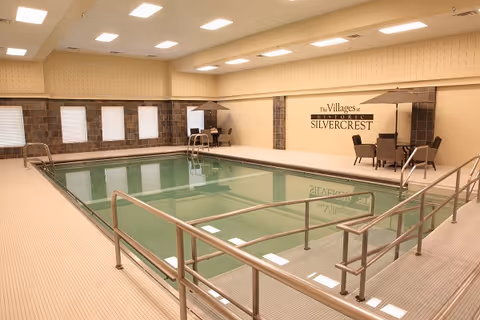 Indoor swimming pool area with handrails and steps leading into the pool. The room has beige tiled flooring and walls with several windows covered by white blinds. There are two seating areas with tables and chairs under umbrellas along the far wall, which displays the sign 'The Villages at Historic Silvercrest'. The ceiling has multiple recessed lights.
