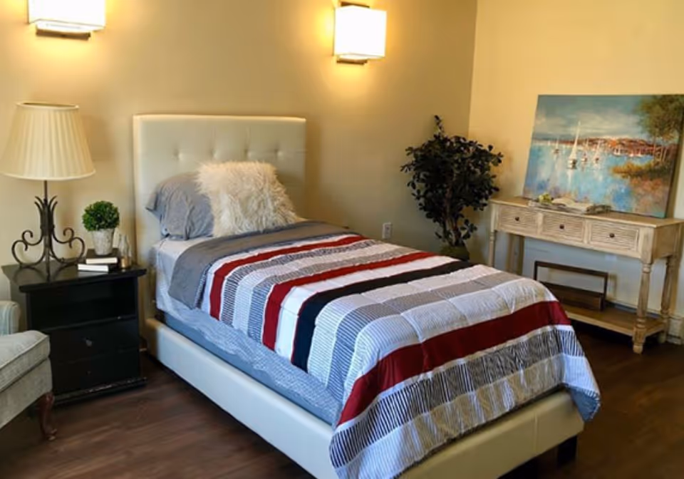 A neatly made single bed with a striped comforter in shades of red, white, black, and gray, placed against a beige wall. The bed has a white tufted headboard and a fluffy white pillow. To the left of the bed is a black nightstand with a decorative lamp, a small plant, and some books. To the right is a wooden console table with three drawers, a painting of a coastal scene resting on it, and a potted plant beside it. The room has wooden flooring and soft lighting from wall-mounted lamps.