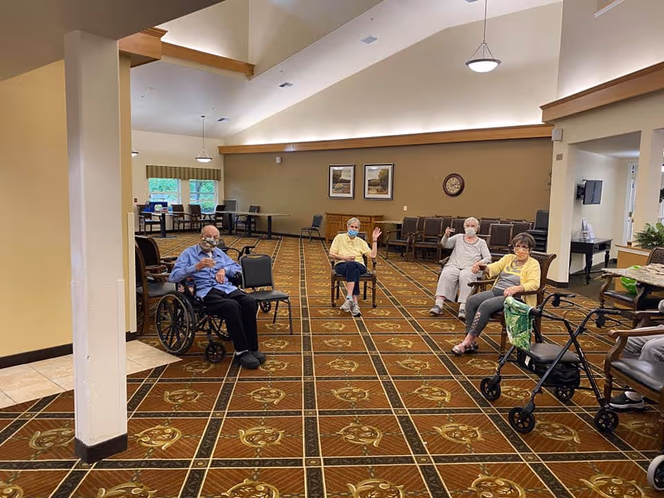 A spacious common area in a senior living facility with four elderly individuals sitting on chairs spaced apart on a patterned carpet. One person is in a wheelchair, and another has a walker nearby. The room has high ceilings with hanging lights, framed pictures on the wall, and several empty chairs and tables in the background.