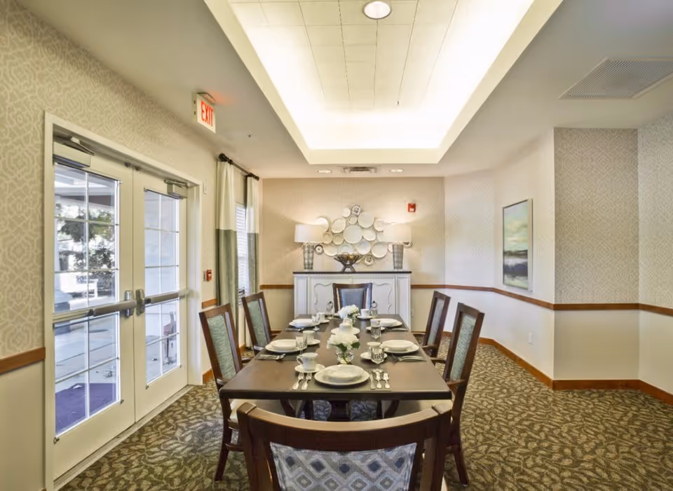 A well-lit dining room with a rectangular wooden table set for six people. The table is arranged with plates, cups, glasses, and silverware, and decorated with small floral centerpieces. The room has patterned carpet, light-colored wallpaper with a subtle design, and a sideboard against the far wall with two lamps and decorative plates mounted above it. There are large glass double doors on the left side letting in natural light.