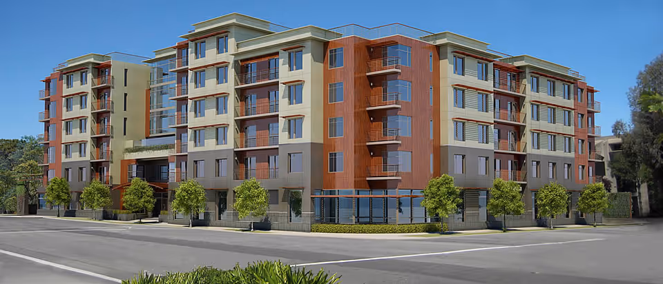 Exterior view of a modern multi-story residential building with balconies and large windows, surrounded by small trees and greenery under a clear blue sky.