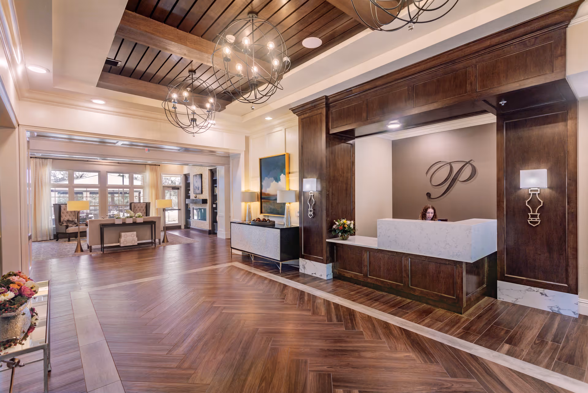 Spacious and elegant reception area of a senior living facility with a wooden front desk, a receptionist seated behind it, decorative wall sconces, and a large letter 'P' on the wall. The room features wooden flooring, modern chandeliers, a seating area with chairs and tables near large windows, and tasteful artwork on the walls.
