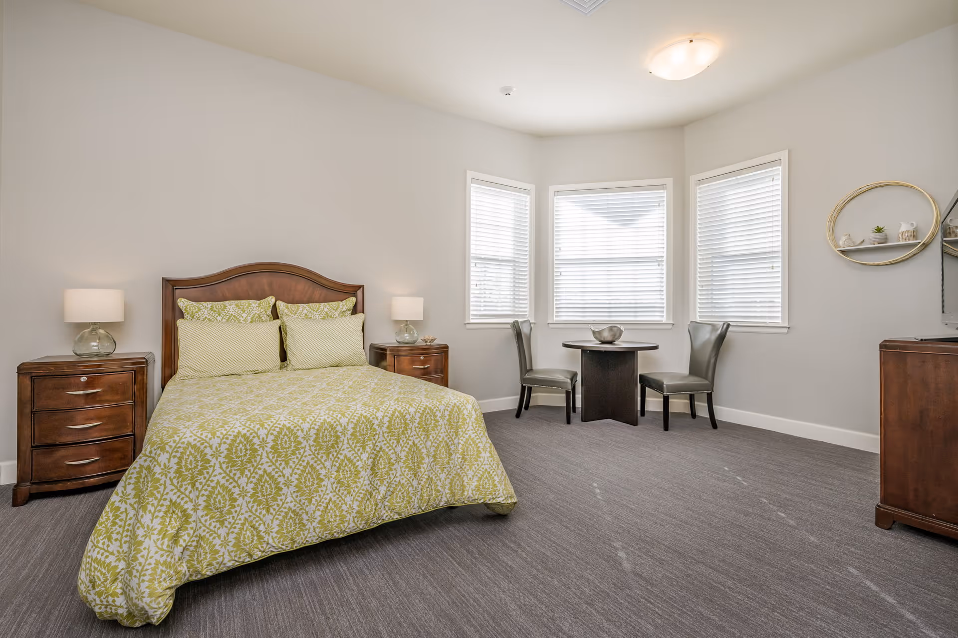 A bedroom with a bed featuring a green and white patterned comforter and matching pillows. There are two wooden nightstands with glass lamps on either side of the bed. Three windows with white blinds are in the corner, next to a small round table with two gray chairs. A wooden dresser and a round wall shelf with small decorative items are also visible.