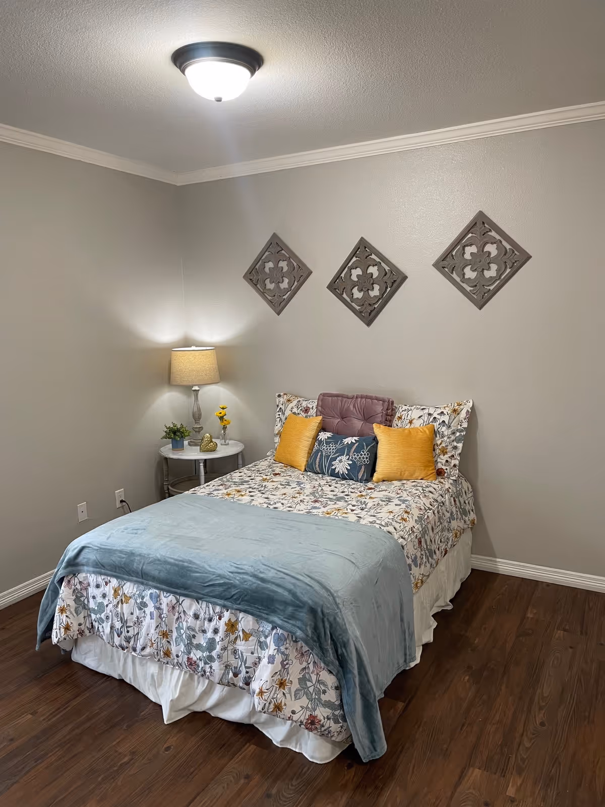 A cozy bedroom with a bed covered in floral bedding, a light blue throw blanket, and several decorative pillows in yellow, purple, and blue. There is a round side table with a lamp, a small plant, and a decorative item. Three square decorative wall hangings are arranged in a row above the bed. The room has wooden flooring and light gray walls.
