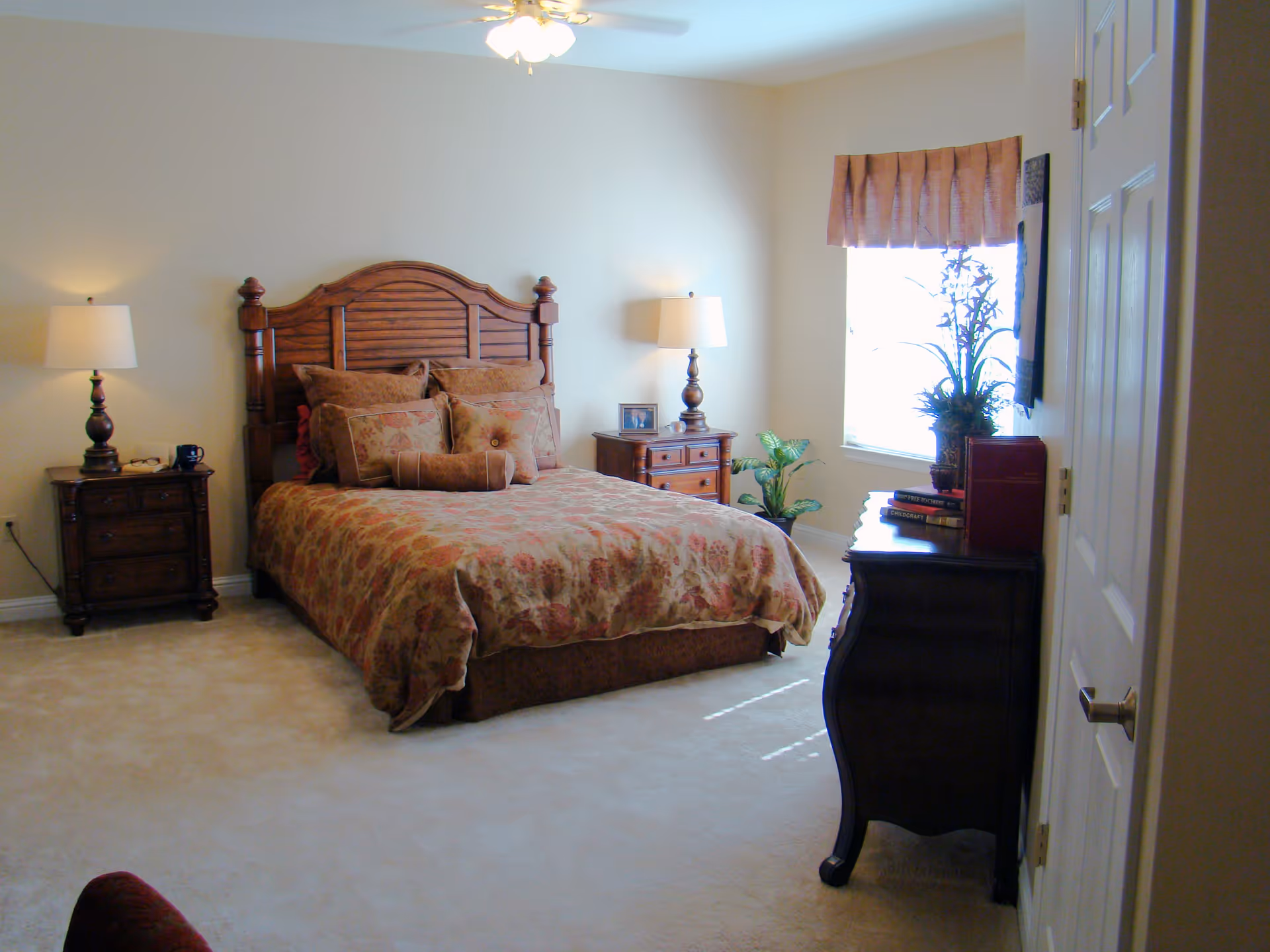A bedroom with a wooden bed frame and floral patterned bedding, two matching nightstands with lamps on either side of the bed, a window with a valance letting in natural light, a dresser with books and a plant on top, and a ceiling fan with lights.