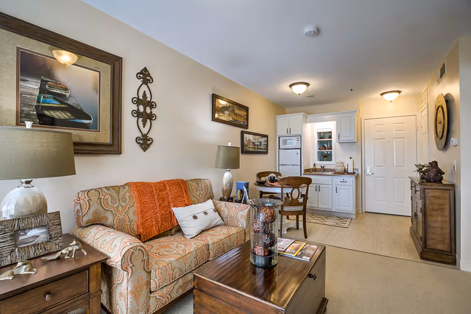 Cozy living area in a retirement community apartment featuring a patterned sofa with an orange throw and a white pillow, a wooden coffee table with decorative items, a side table with a lamp and picture frame, and a small dining table with two chairs. The background shows a compact kitchen with white cabinets, a refrigerator, microwave, and a white entrance door. The walls are decorated with framed artwork and a decorative wall piece.
