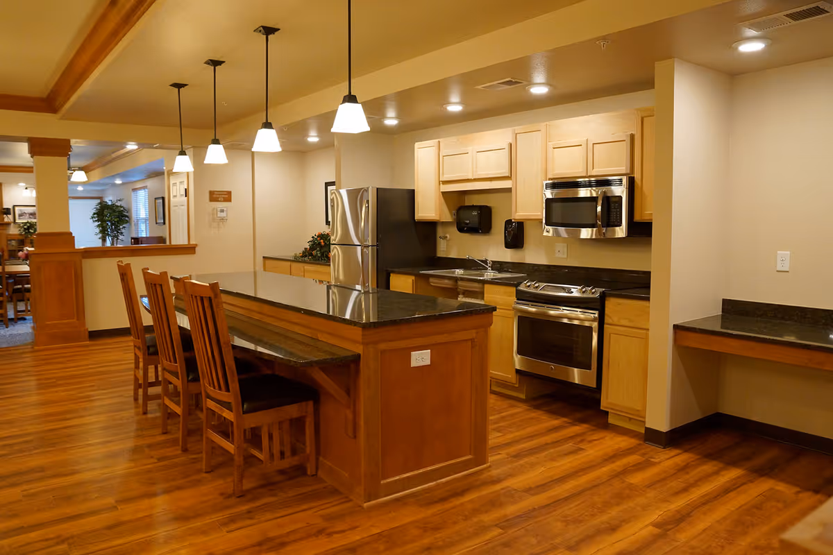 A modern kitchen area with wooden cabinets, stainless steel refrigerator, oven, and microwave. There is a large island with a dark countertop and four wooden chairs. Pendant lights hang above the island, and the floor is wood. The kitchen opens into a dining or common area with additional seating and plants.